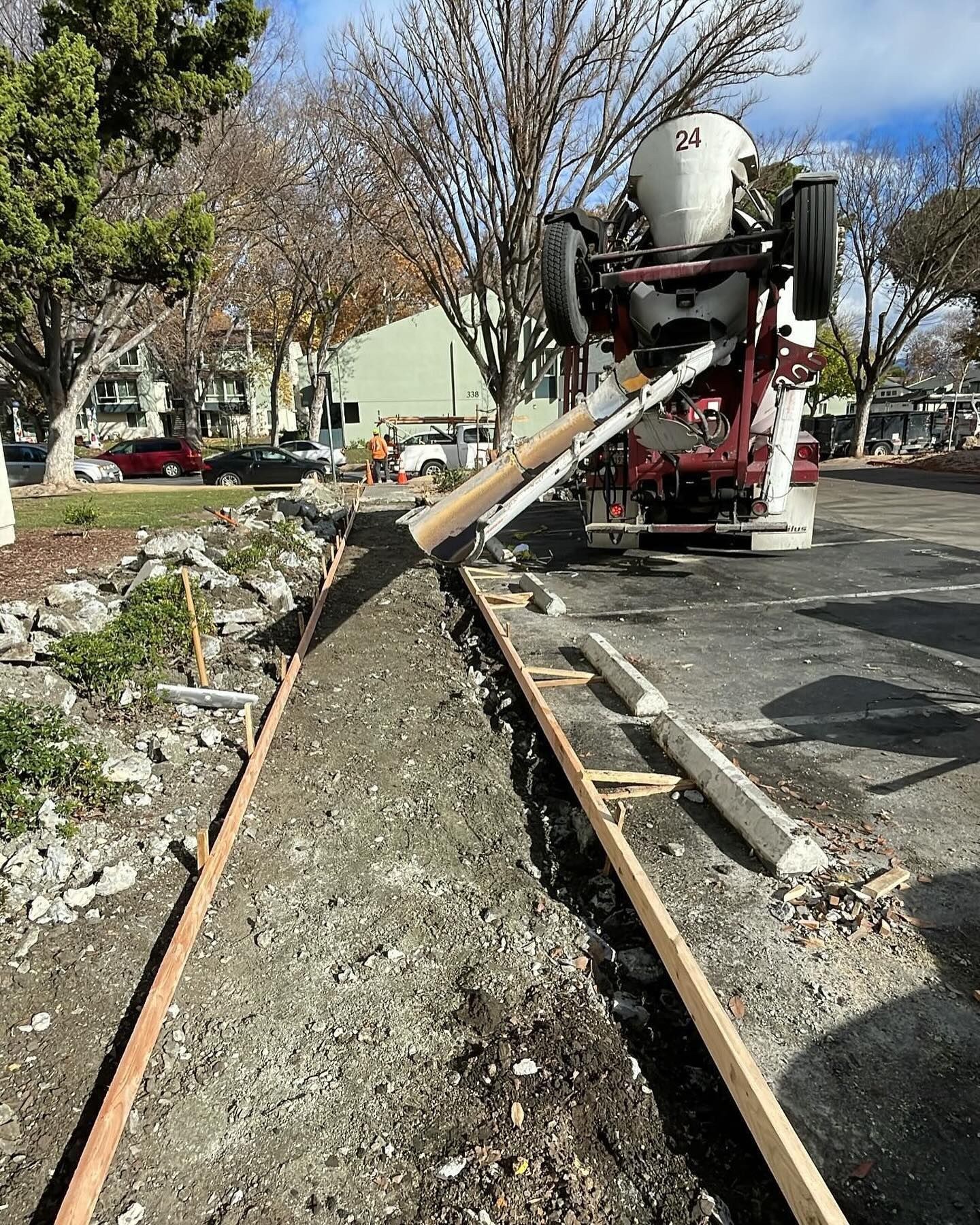 A concrete mixer truck pours wet cement into a framed-out trench for a new sidewalk near a parking lot.