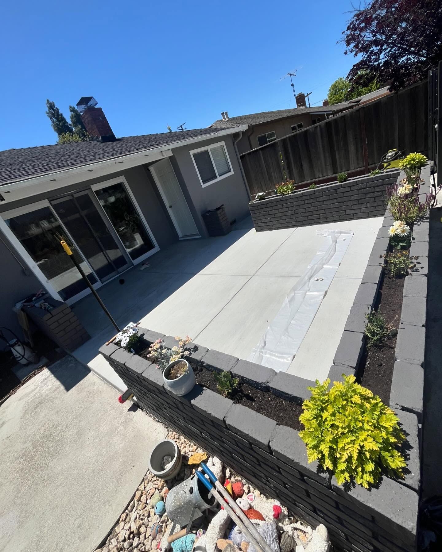 A patio with gray concrete pavers, dark gray stacked stone planter walls, and a new bright green plant in a sunny backyard.