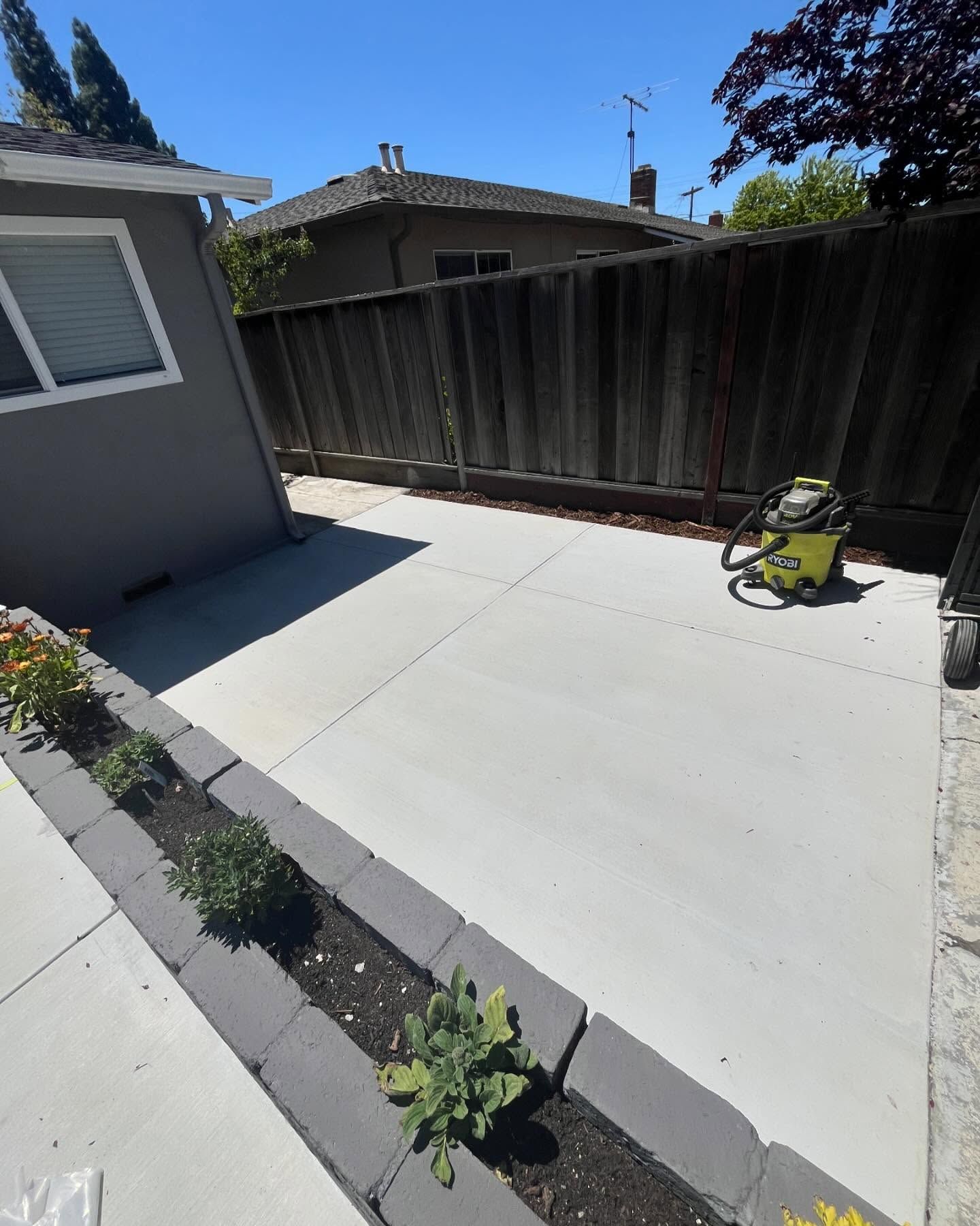 A gray concrete patio beside a house, featuring a long, rectangular planter box edged with dark gray bricks.