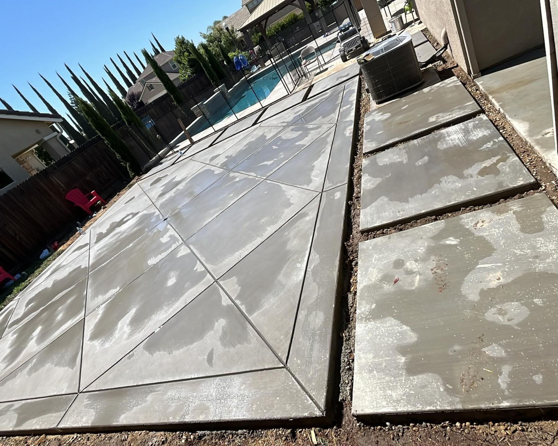 A high-angle view of freshly poured concrete patio slabs with geometric scoring, adjacent to a backyard pool and fencing.