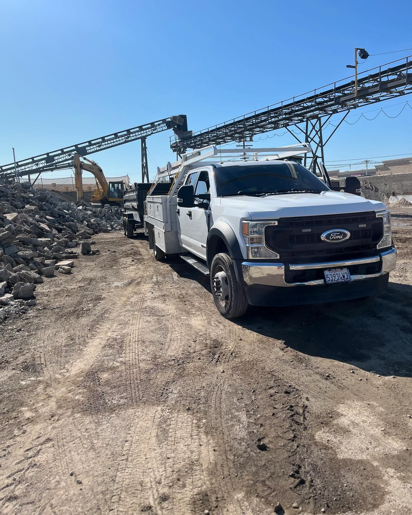 A white service truck parked on a dirt lot at a construction site with heavy machinery and industrial equipment nearby.