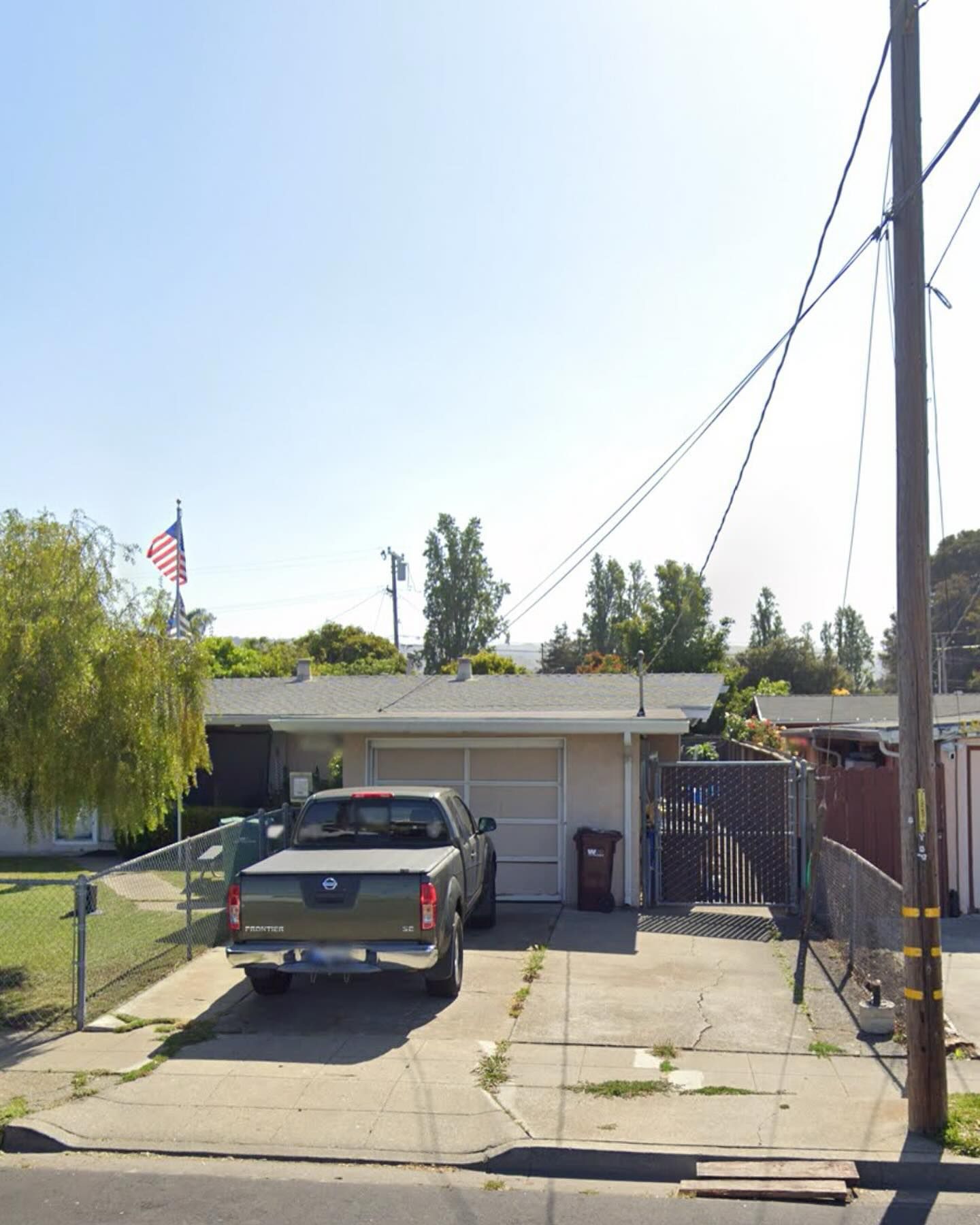 A single-story house with a driveway, a parked gray pickup truck, a chain-link fence, and an American flag.