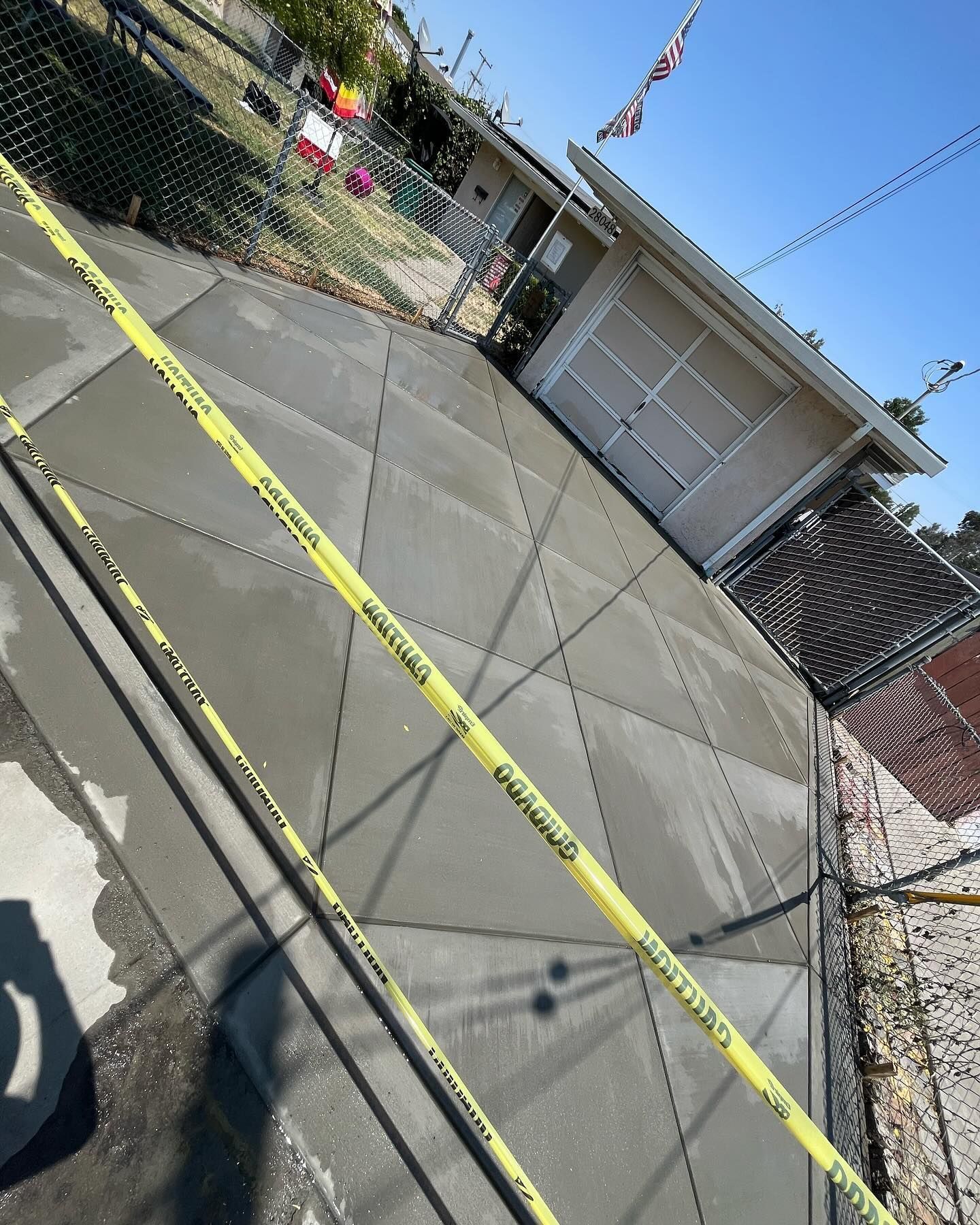 A freshly poured concrete driveway sectioned into a diamond pattern, blocked off by yellow caution tape under a clear sky.