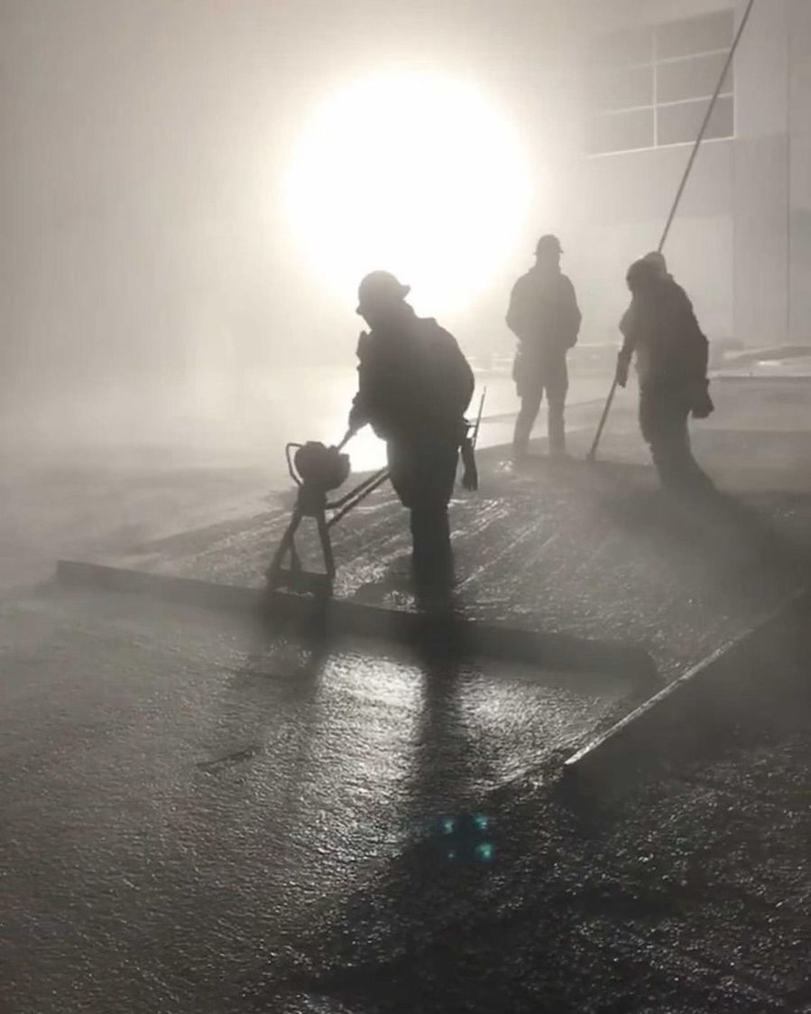 Three workers in hard hats use equipment to smooth wet concrete outdoors on a foggy, brightly lit day.