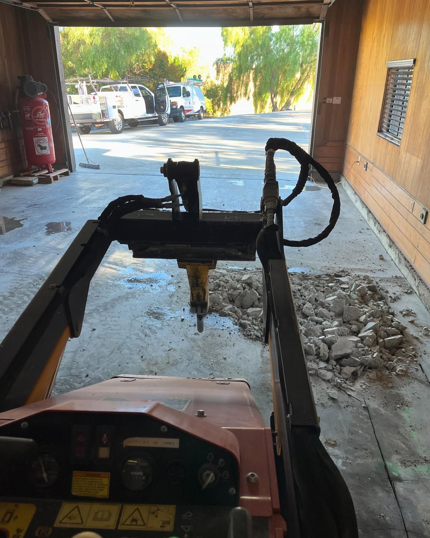 View from a skid steer inside a garage, featuring a hydraulic concrete breaker over a pile of broken concrete rubble.