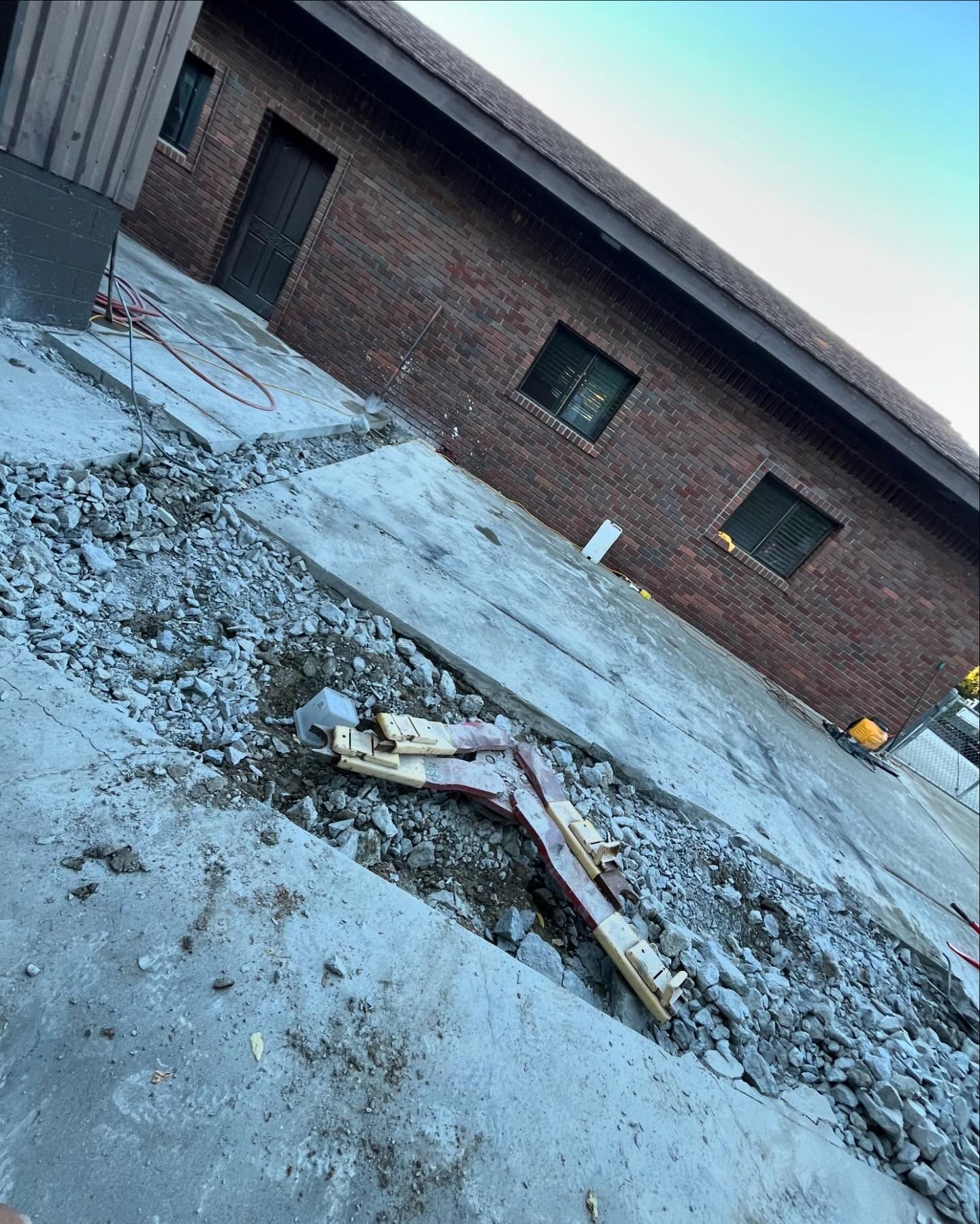 A construction site shows a trench cut through a concrete driveway in front of a brick building with several windows.