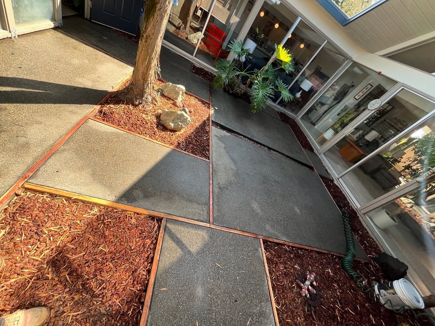 A paved patio with geometric concrete slabs and wood borders, surrounding a tree and garden beds with reddish-brown mulch.