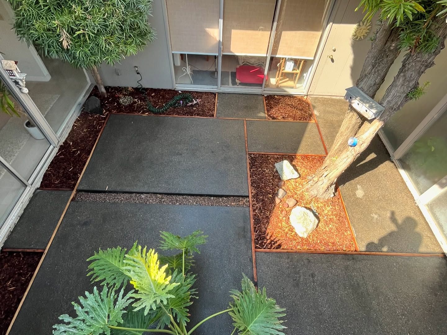 A modern courtyard with dark gray rectangular pavers, reddish mulch beds, a small tree, and a green potted plant.