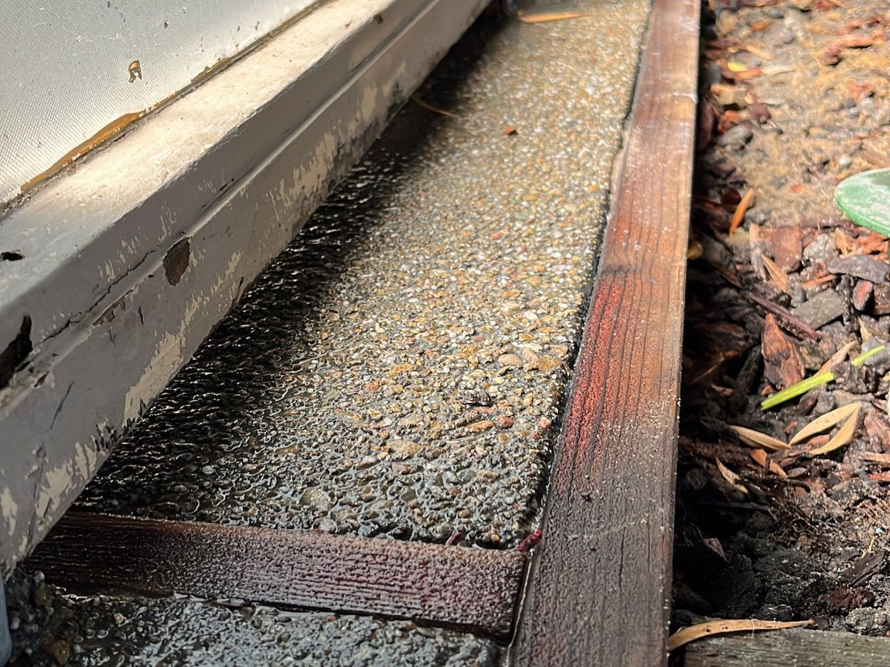 A close-up view of a wet, exposed aggregate concrete strip between a grey metal frame and a wooden border in a garden.