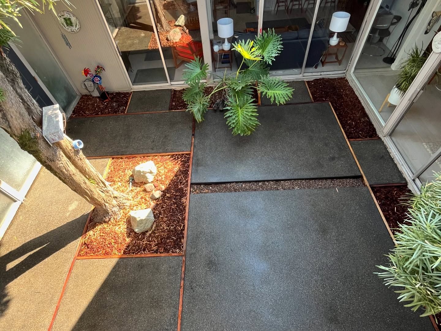 An overhead view of a courtyard with dark concrete pavers, reddish mulch, white rocks, and a leafy green plant.