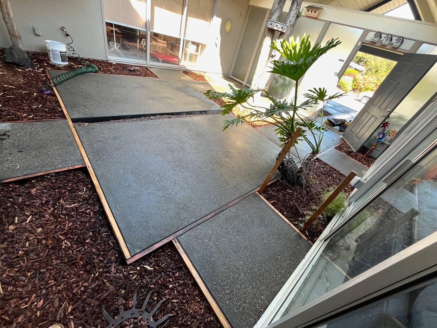 A recently poured concrete patio featuring large gray pavers set in dark wood mulch, situated outside a glass door.