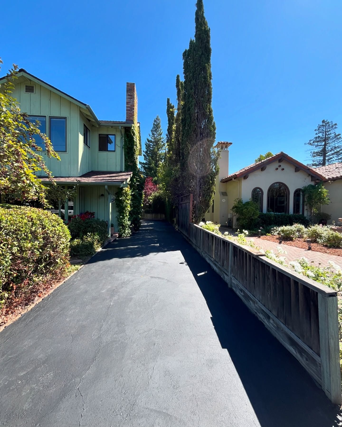 A paved driveway leads between a green two-story house on the left and a stucco home on the right under a clear blue sky.