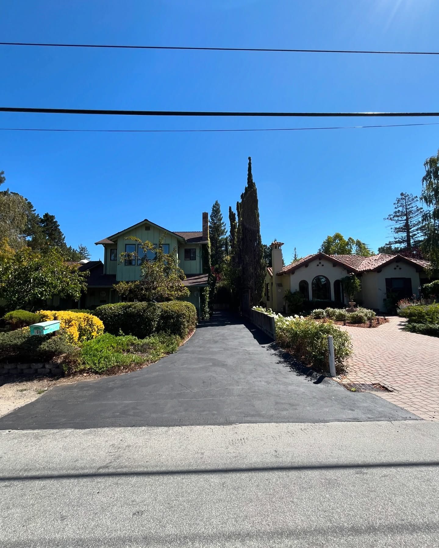 A paved driveway leads toward two houses on a sunny day, with a gravel path to the right and surrounding greenery.
