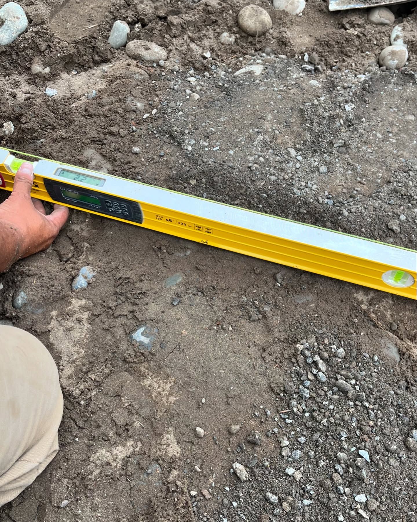 A person holds a yellow digital level against rough, uneven dirt ground to measure its incline.