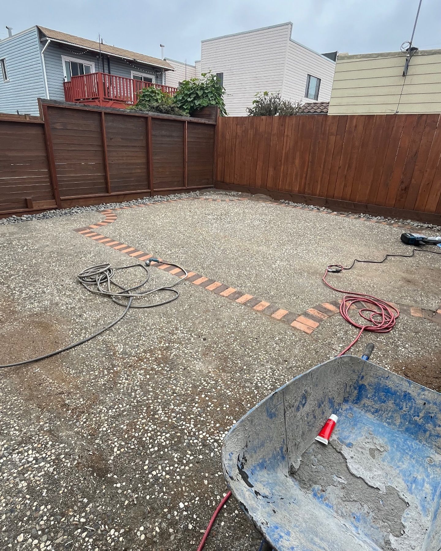 A backyard renovation in progress, featuring a gravel patio area with a brick outline, a wooden fence, and a wheelbarrow.