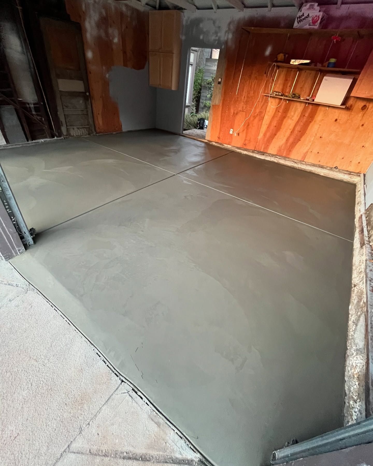 A freshly poured, smooth gray concrete floor in a garage with wooden walls and shelving units in the background.