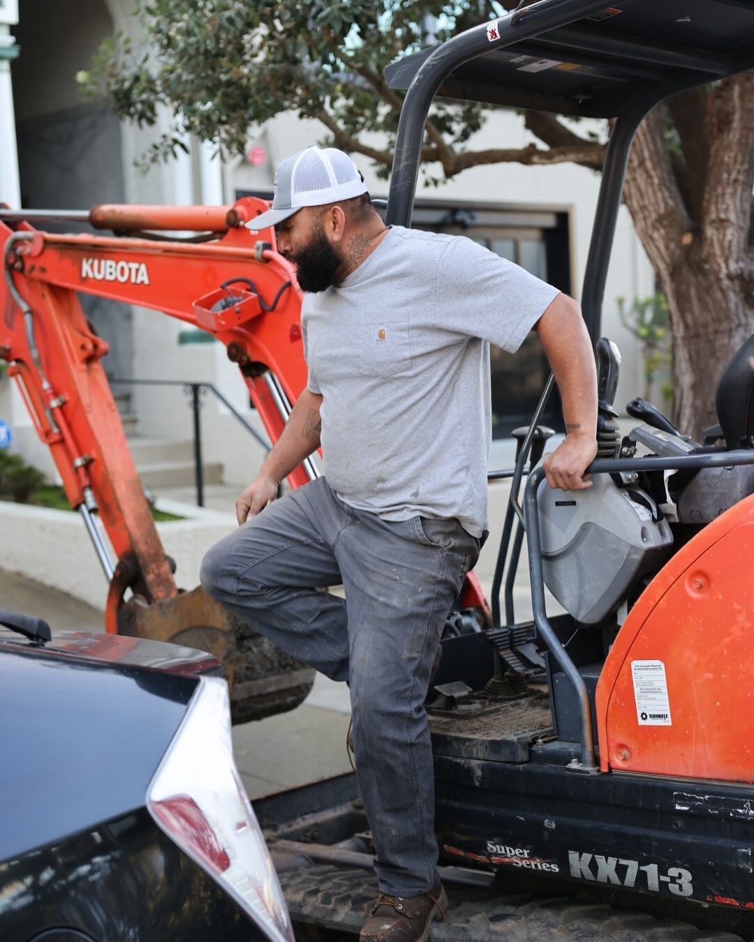 A person in a gray shirt and white cap steps out of an orange Kubota mini excavator parked near a car.