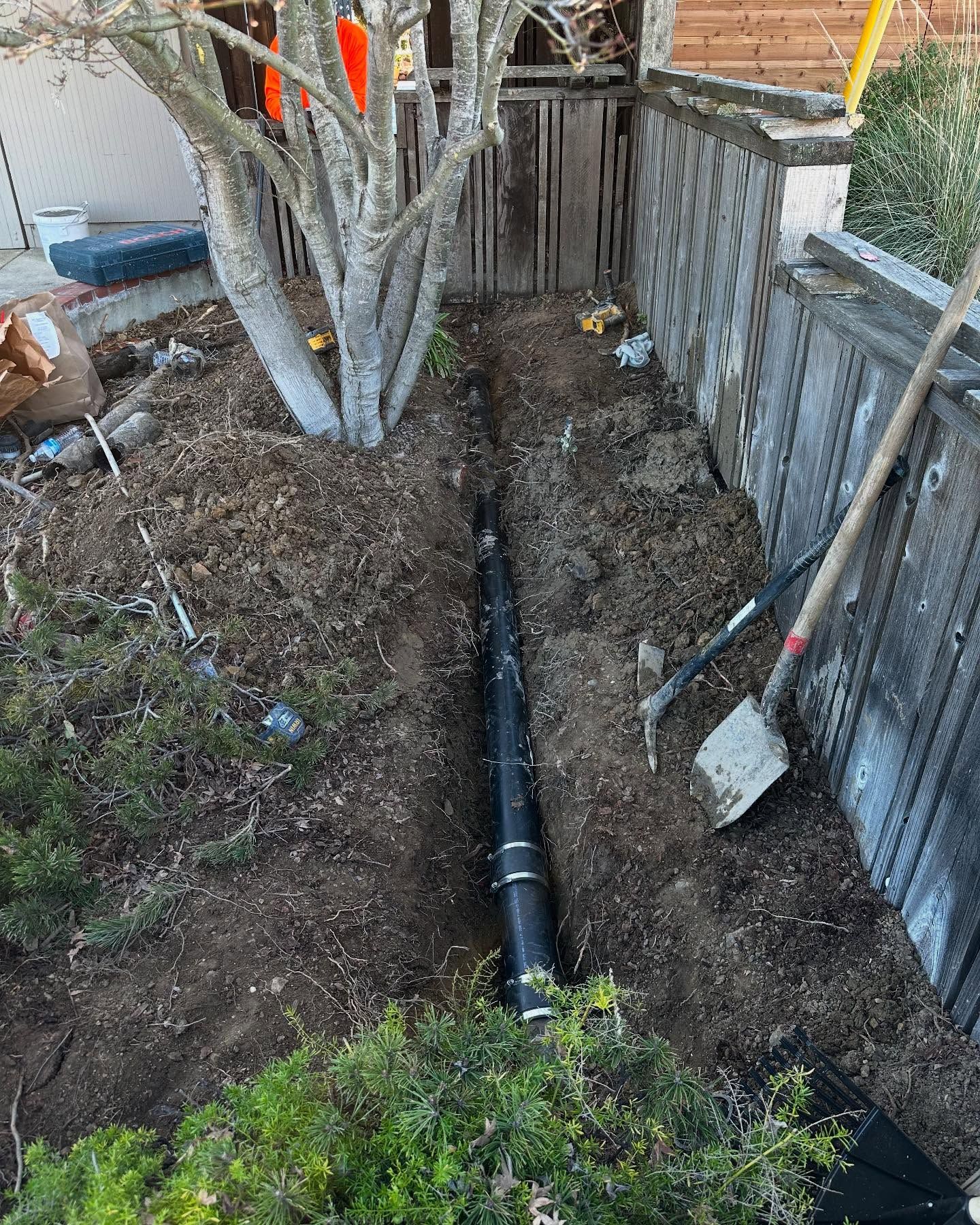 A black drainage pipe installed in a narrow trench next to a wooden fence and a tree.