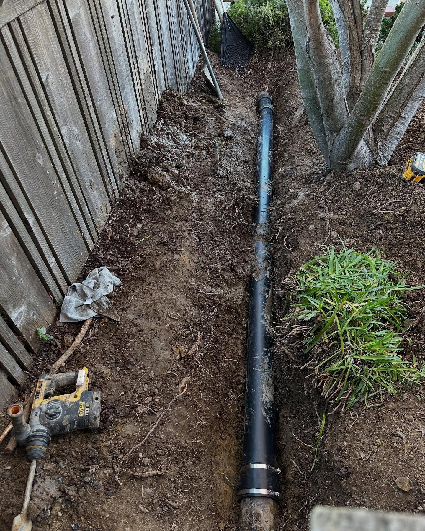 A black plastic drainage pipe sits in a narrow dirt trench next to a wooden fence.