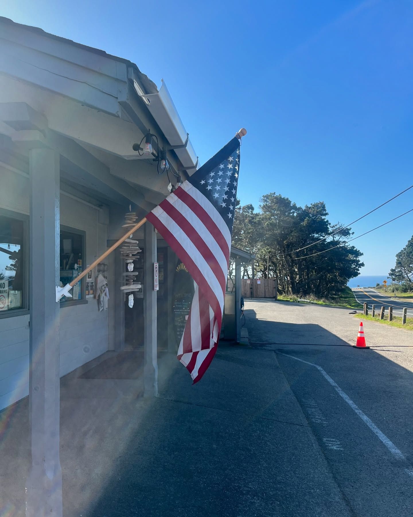 An American flag hangs from the porch of a building next to a paved driveway and trees under a clear blue sky.