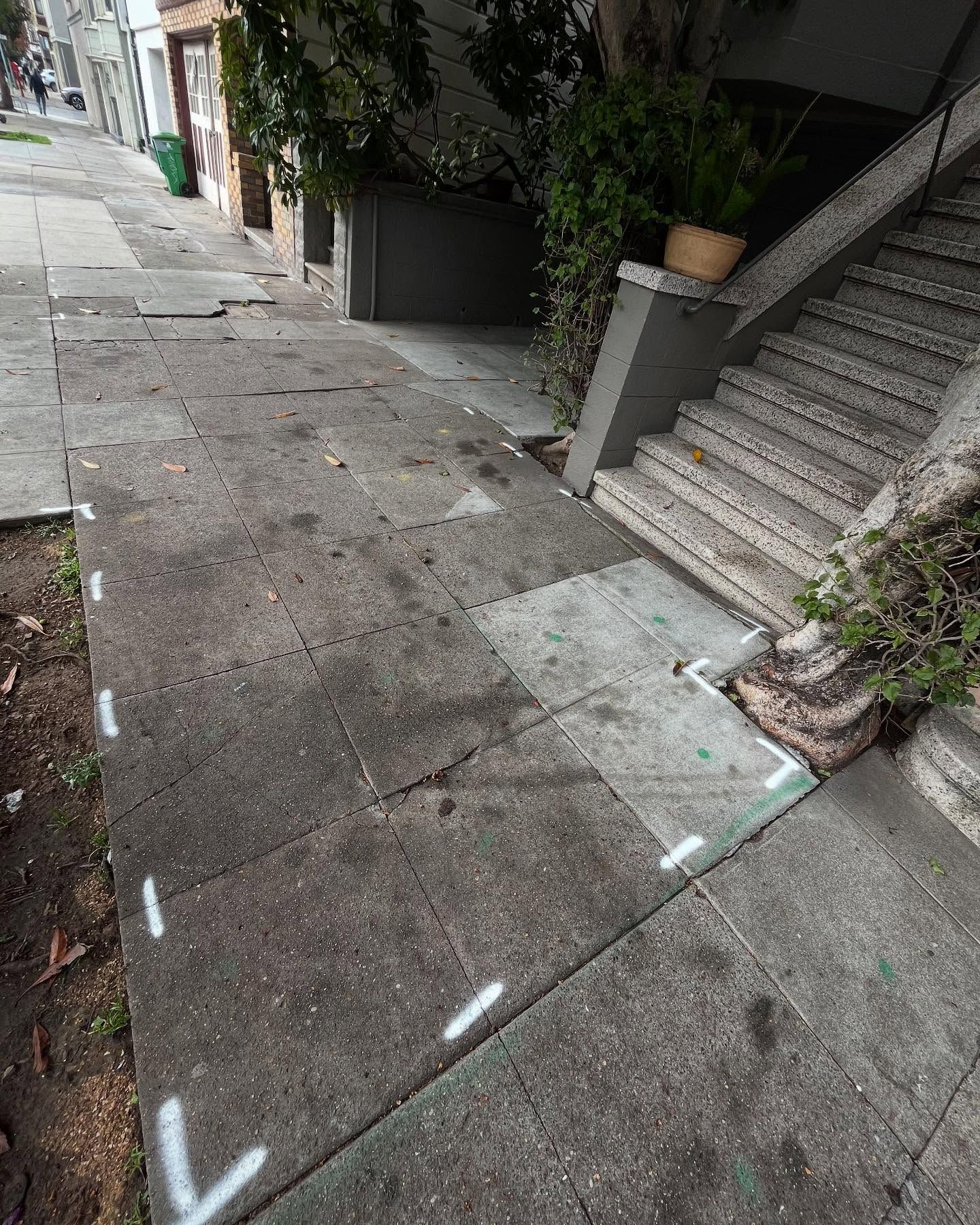 A sidewalk with white construction markings outlining sections of concrete near a building entrance with stone stairs.
