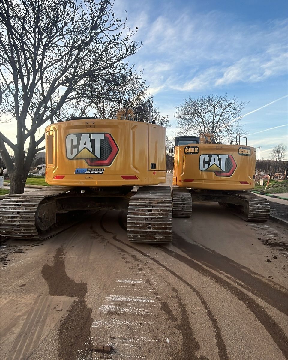 Two yellow Caterpillar excavators parked side-by-side on a dirt road next to a tree under a blue sky.