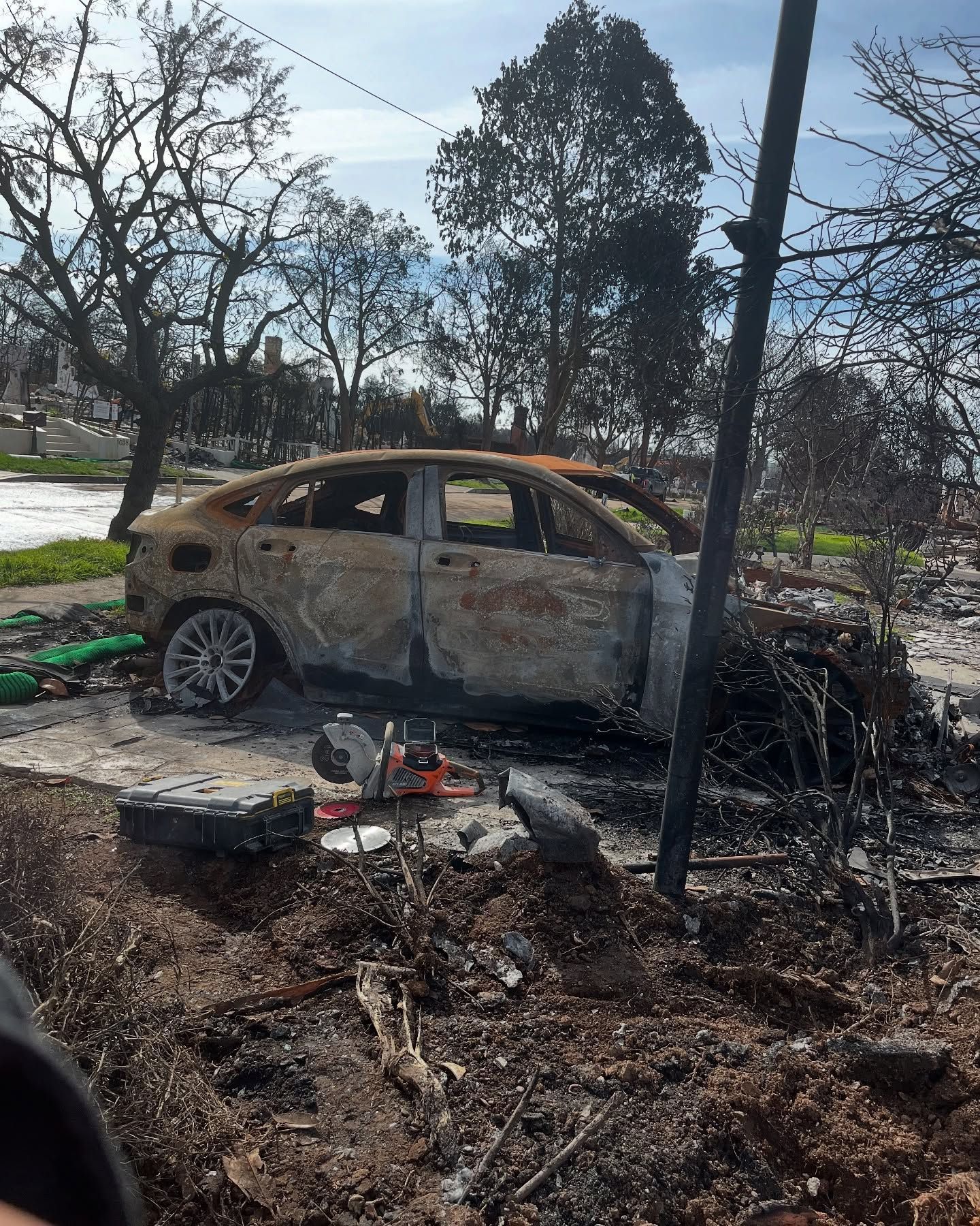 A burned-out sedan sits in a landscape of charred debris and scorched trees under a bright, clear sky.