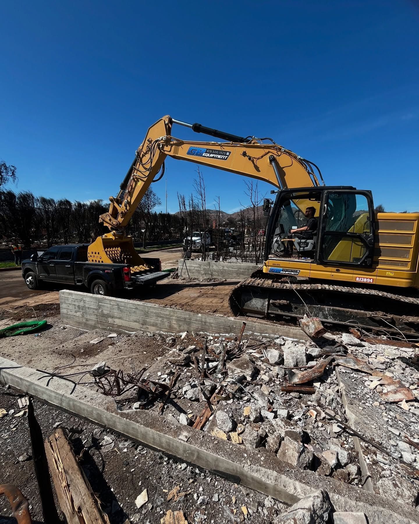 A yellow excavator is parked next to a black pickup truck at a demolition or disaster cleanup site under a clear sky.