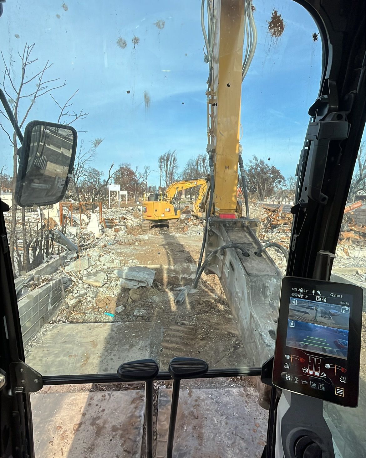 View from inside an excavator cab showing another yellow excavator operating in a debris-filled area after a fire.