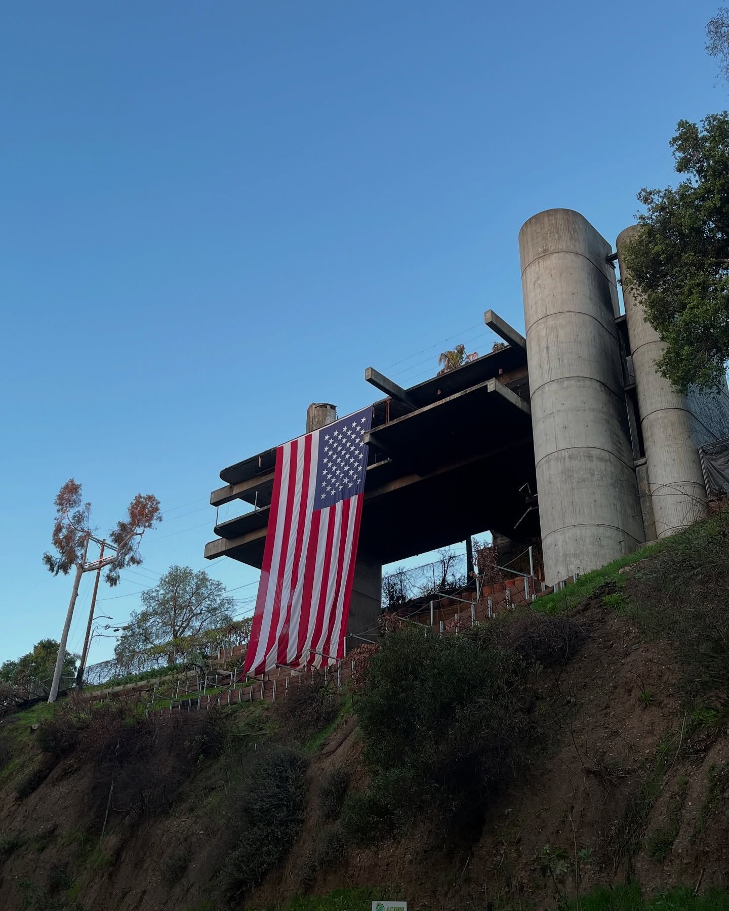 An American flag hangs from the concrete skeleton of a partially demolished building atop a hillside against a blue sky.