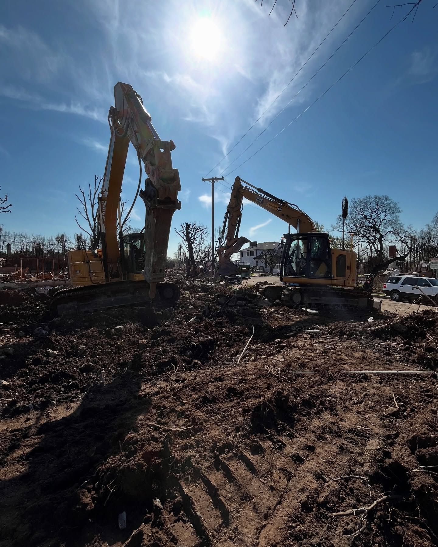 Two yellow excavators work in a debris-filled field under a bright sun.
