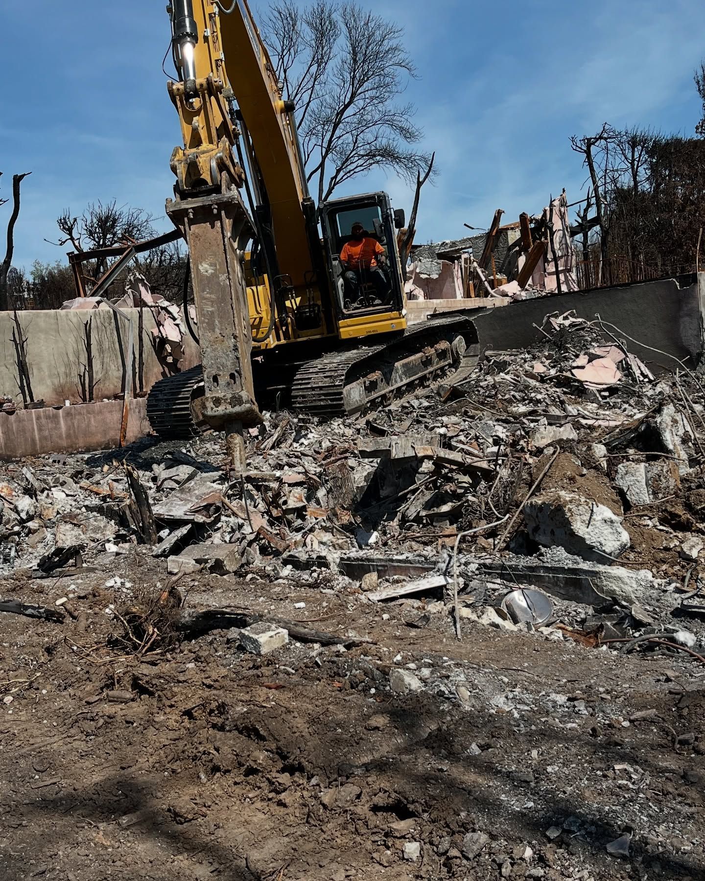 A yellow excavator with a hydraulic breaker attachment works on a pile of concrete rubble at a demolition site.