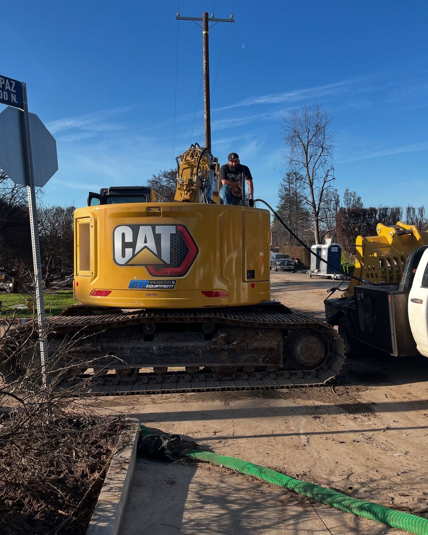 A worker performs maintenance on the engine of a yellow Caterpillar excavator parked on a dirt lot under a blue sky.