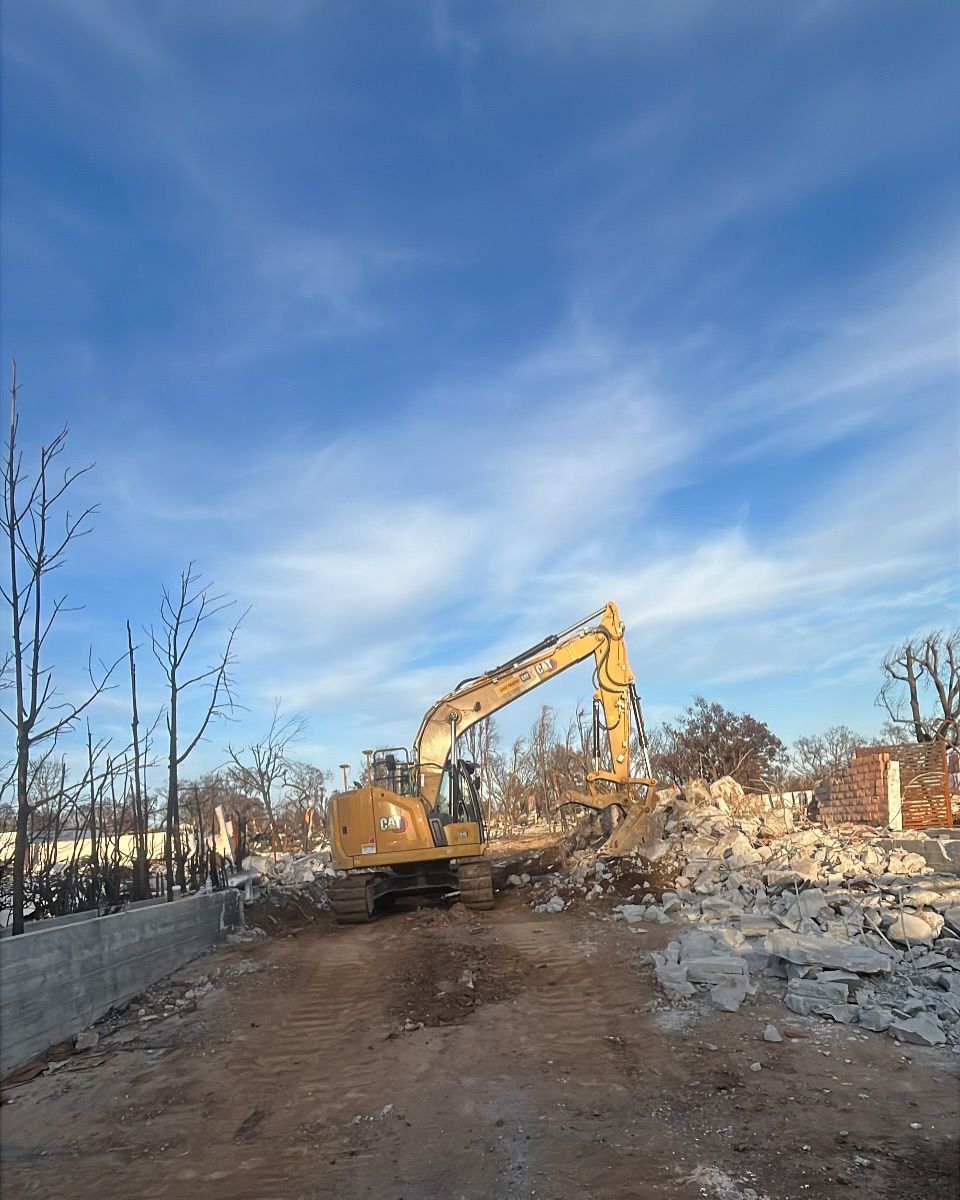 A yellow excavator works in a field of construction rubble against a clear blue sky.