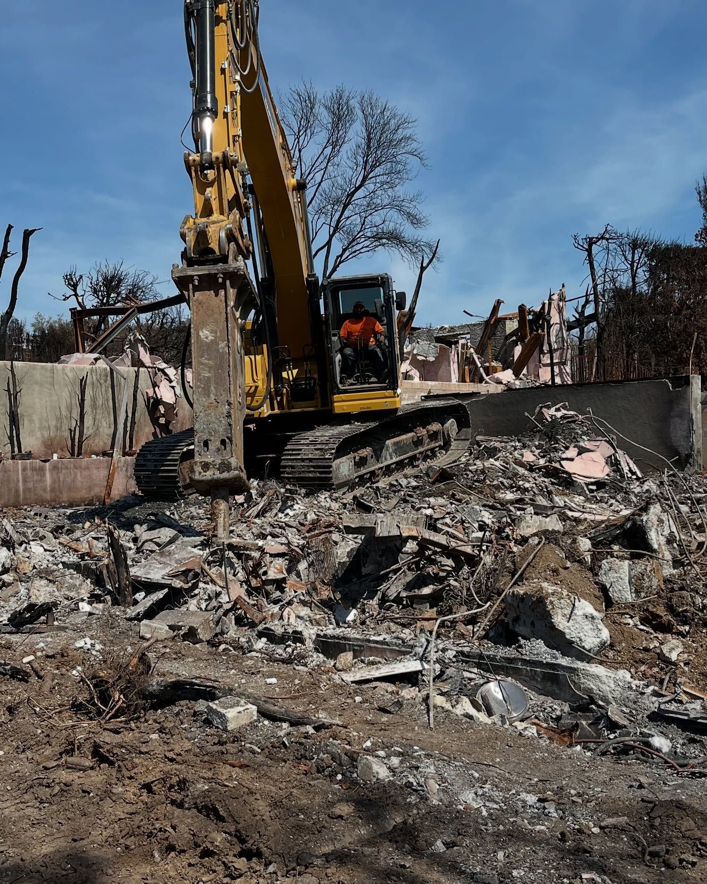 A yellow excavator with a hydraulic breaker attachment works on a pile of debris at a demolition or disaster site.