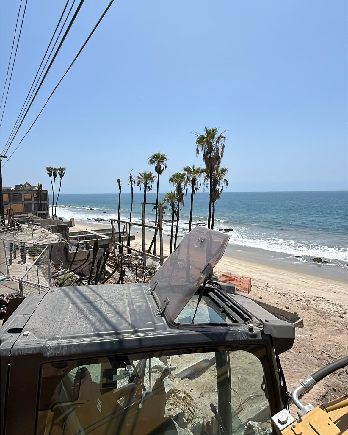 A construction vehicle overlooking a beach with palm trees and a building foundation under a clear, bright blue sky.