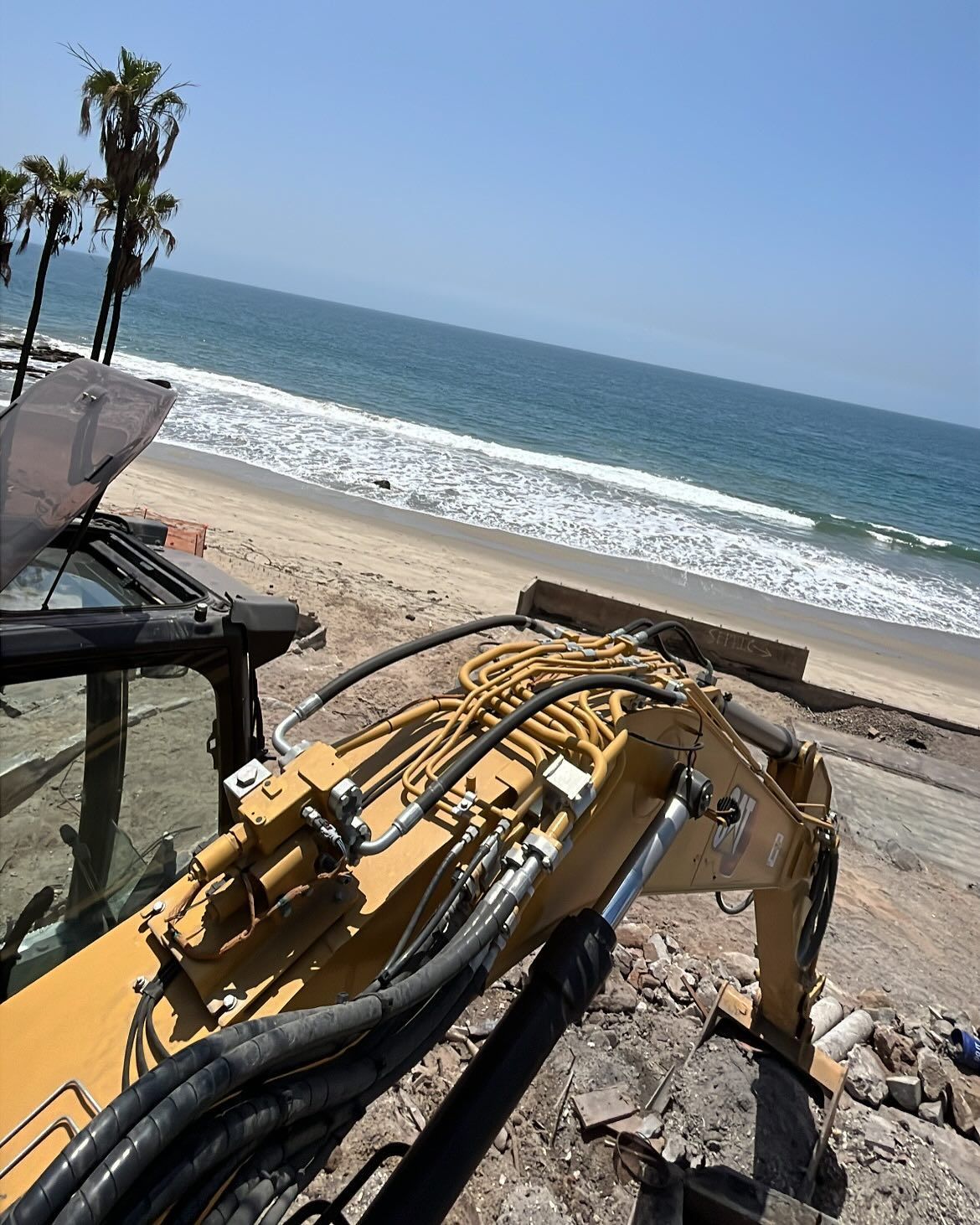 A yellow excavator arm overlooking a sandy beach and ocean under a clear blue sky.