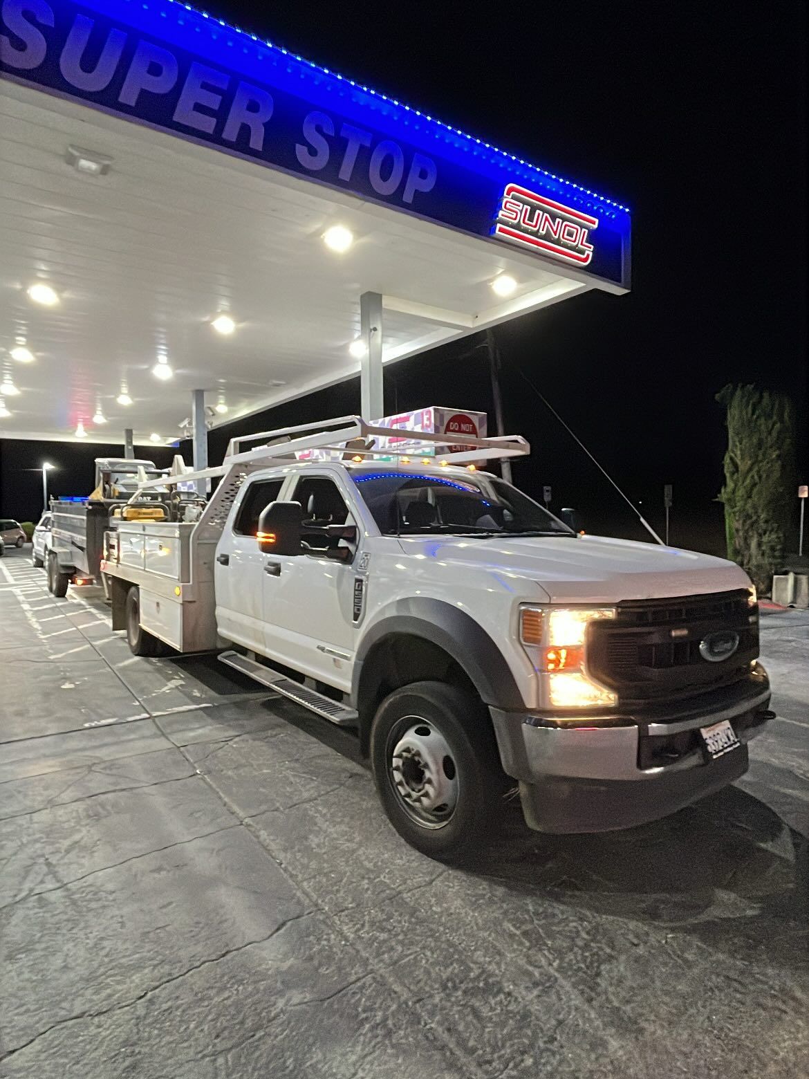 A white service truck with a ladder rack and utility bed parked under a lighted Super Stop gas station canopy at night.