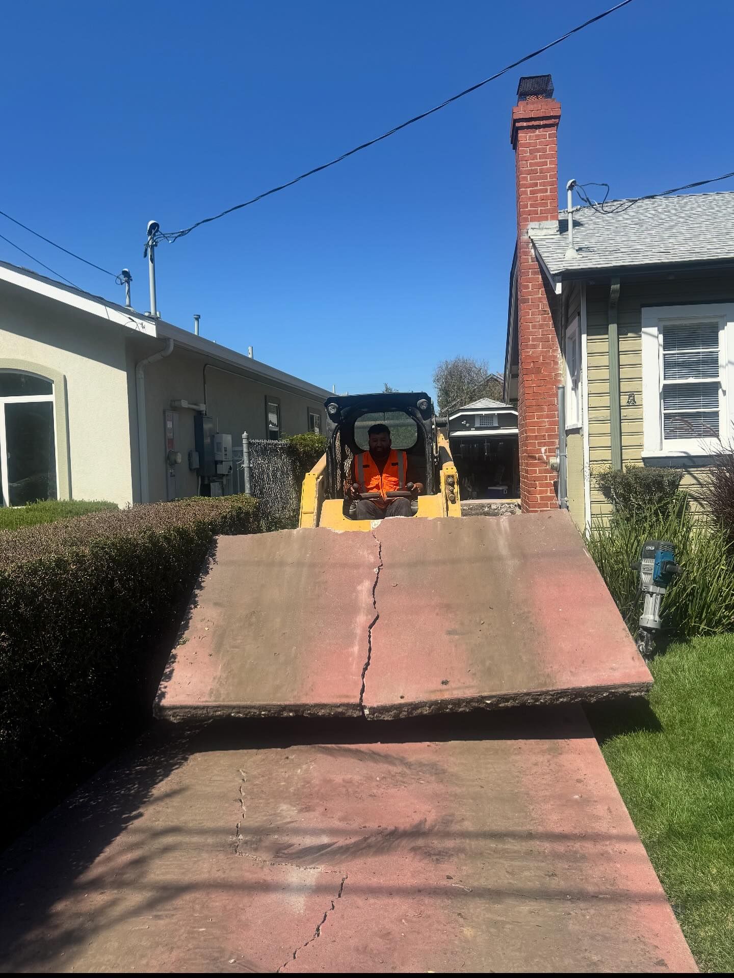 A person in an orange safety vest operates a yellow skid steer to lift a large slab of concrete from a residential driveway.