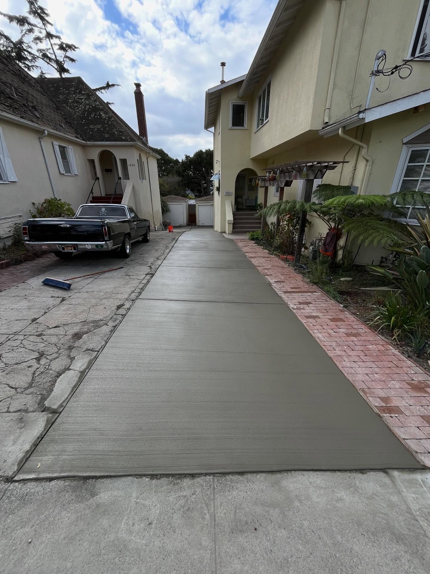 A freshly poured concrete driveway lane between two houses, bordered by an older, cracked driveway and brick pavers.