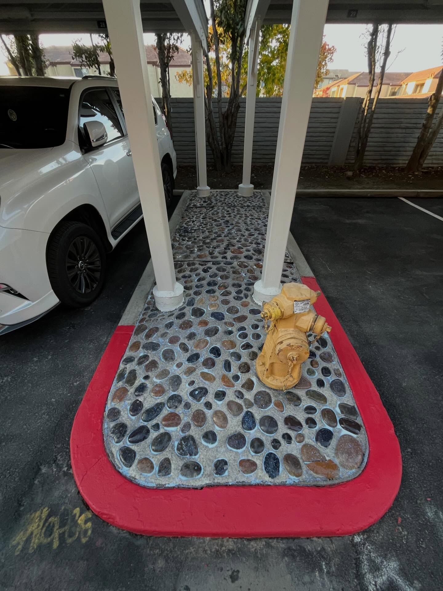 A yellow fire hydrant sits on a pebble-filled island with red-painted curbs between parking spots under a carport.