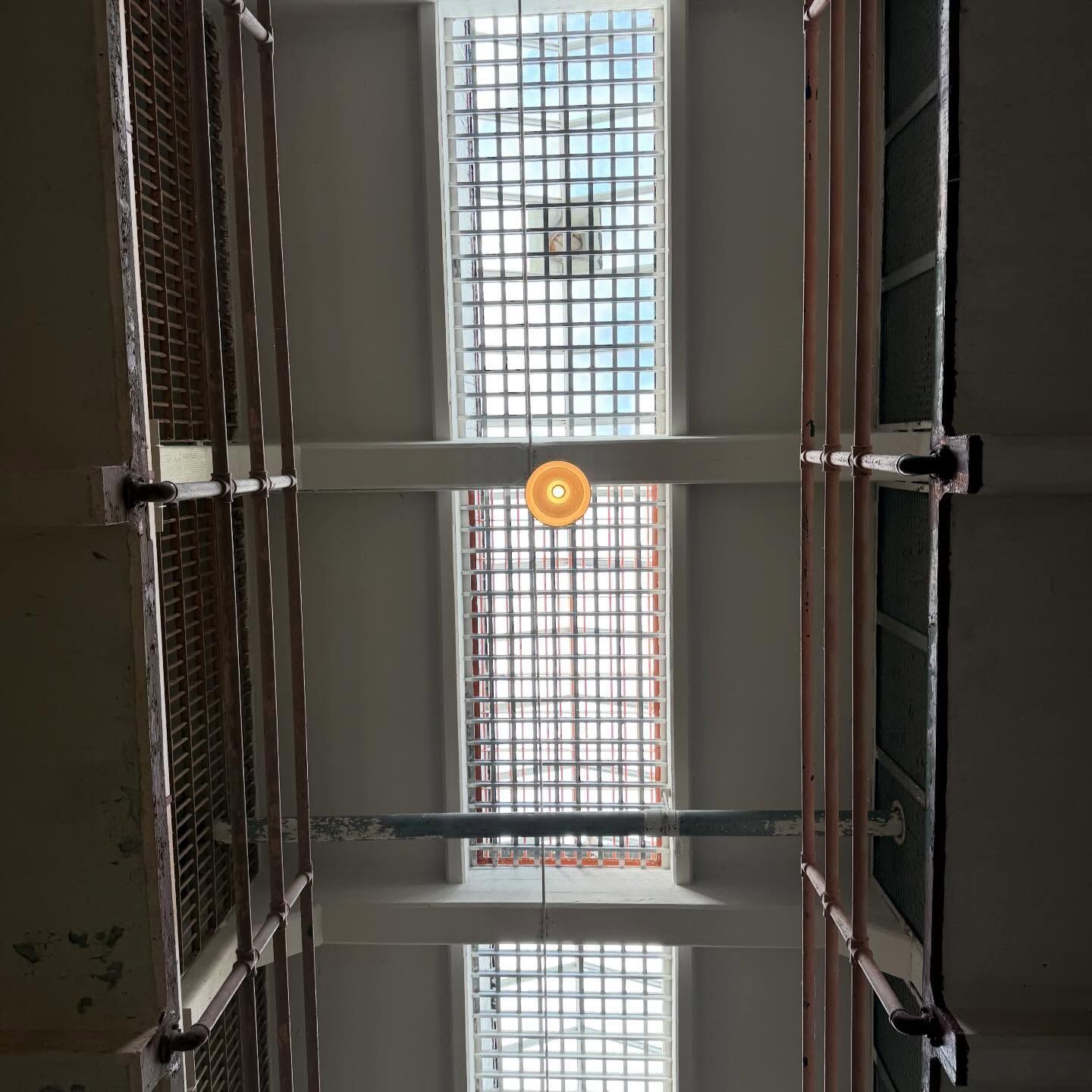 Looking straight up at a skylight with a white grid frame, bisected by concrete beams and a single hanging round light.