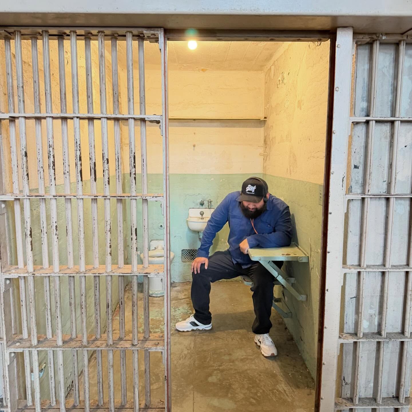 A person in a blue shirt and baseball cap sits on a small bench inside a cramped, rustic prison cell.