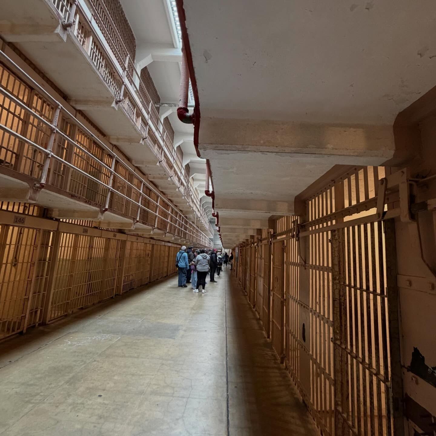 A long, narrow cell block corridor at Alcatraz, with metal barred cells on both sides and a group of visitors walking.