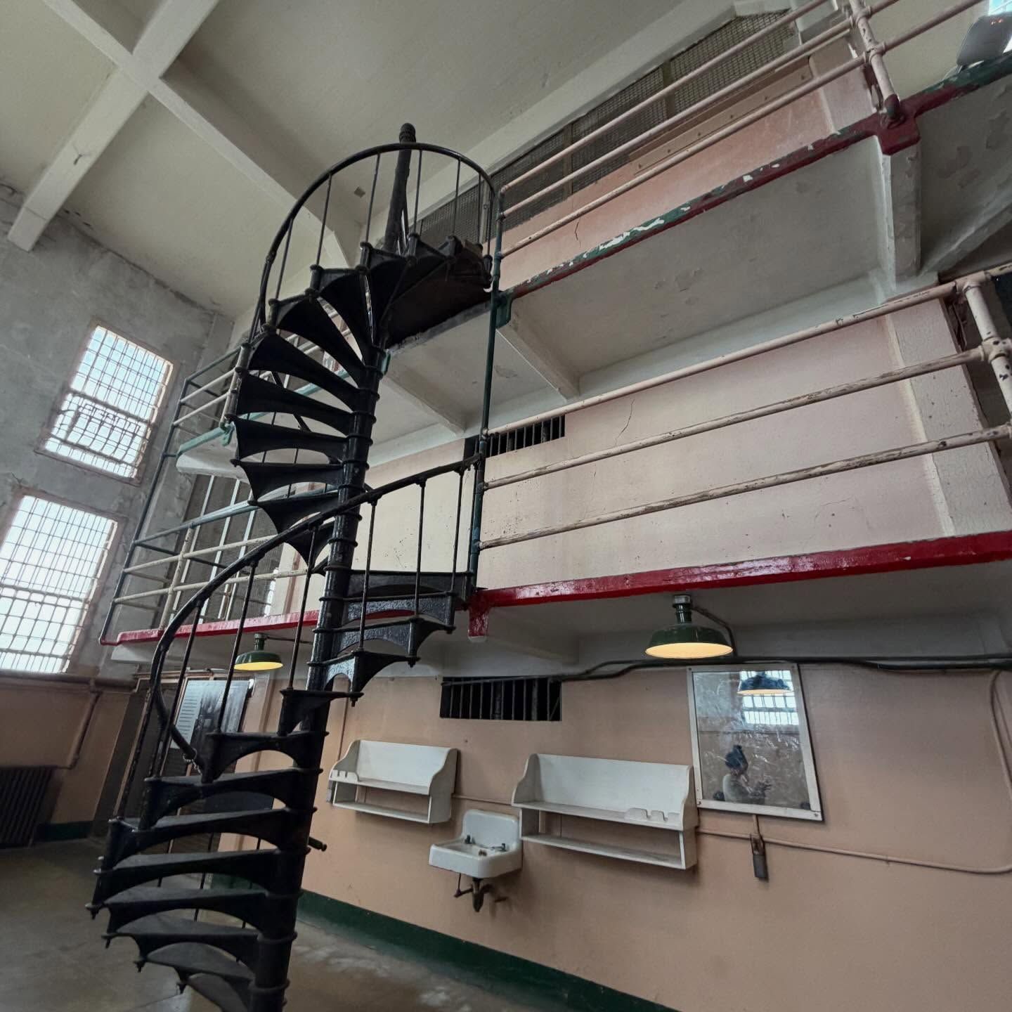 A black spiral staircase stands in a multi-level prison block with barred windows, metal railings, and wall-mounted sinks.