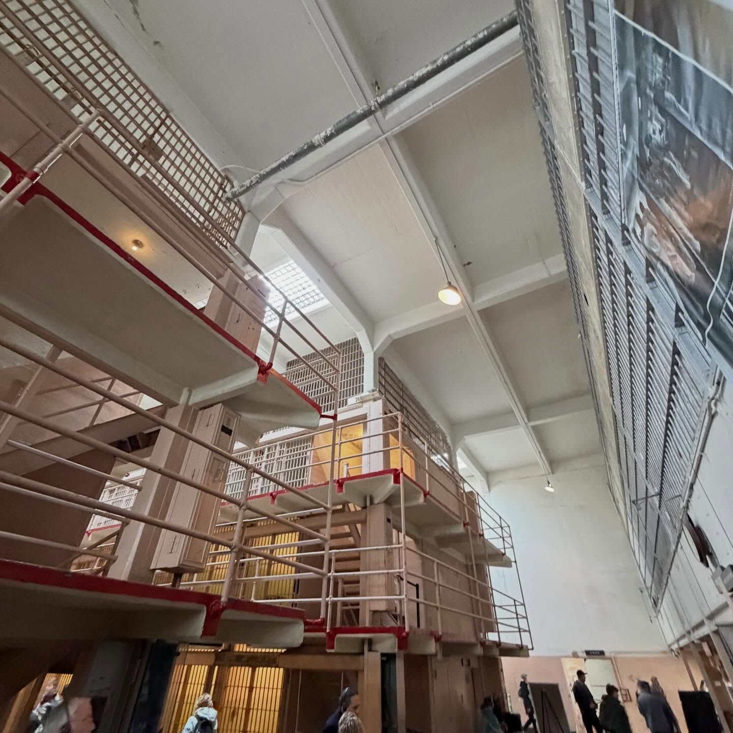 Low-angle view of a multi-story prison block with metal catwalks, mesh railings, and a high ceiling with skylights.