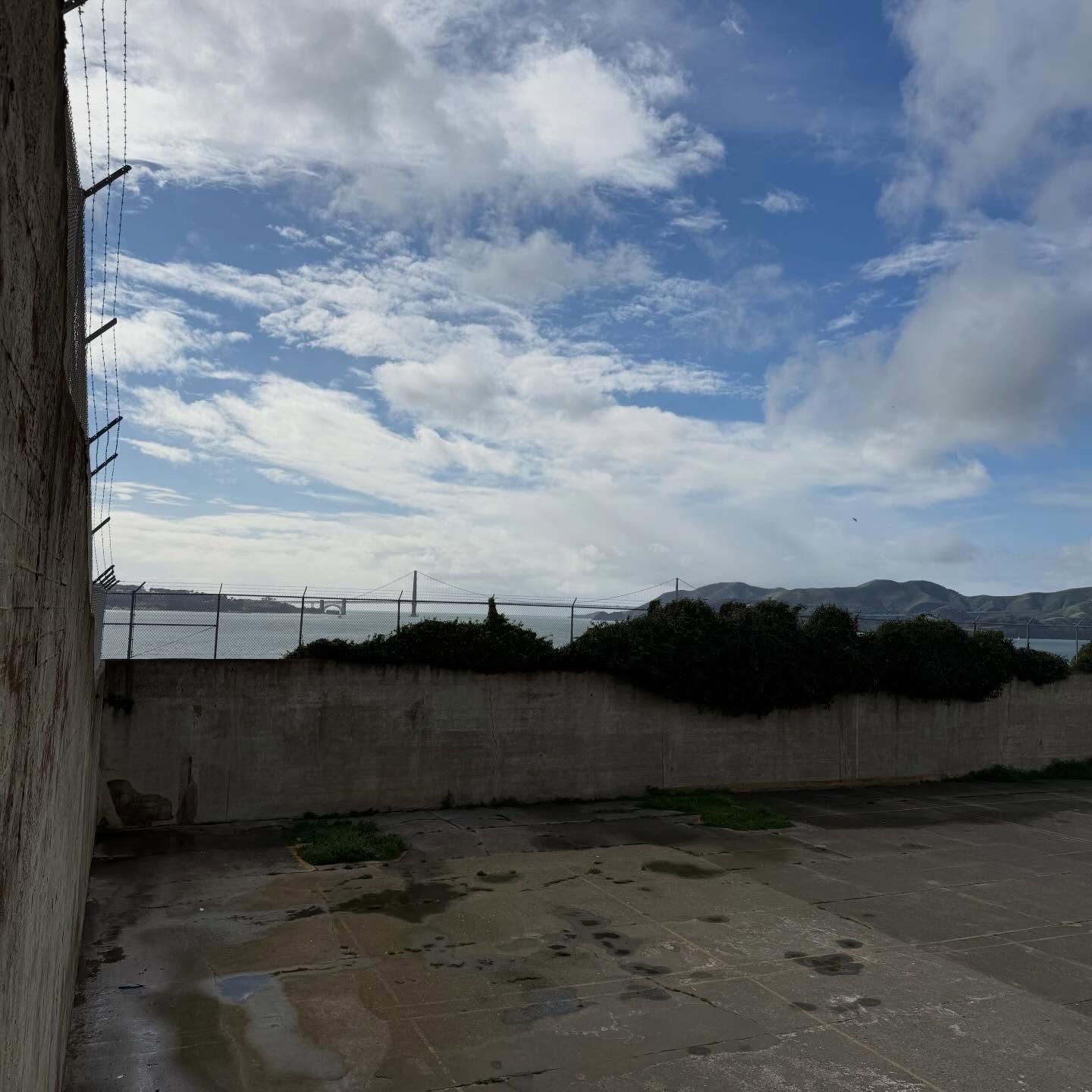 A concrete yard enclosed by a low wall with a fence, looking out toward a distant bridge and hills under a cloudy sky.