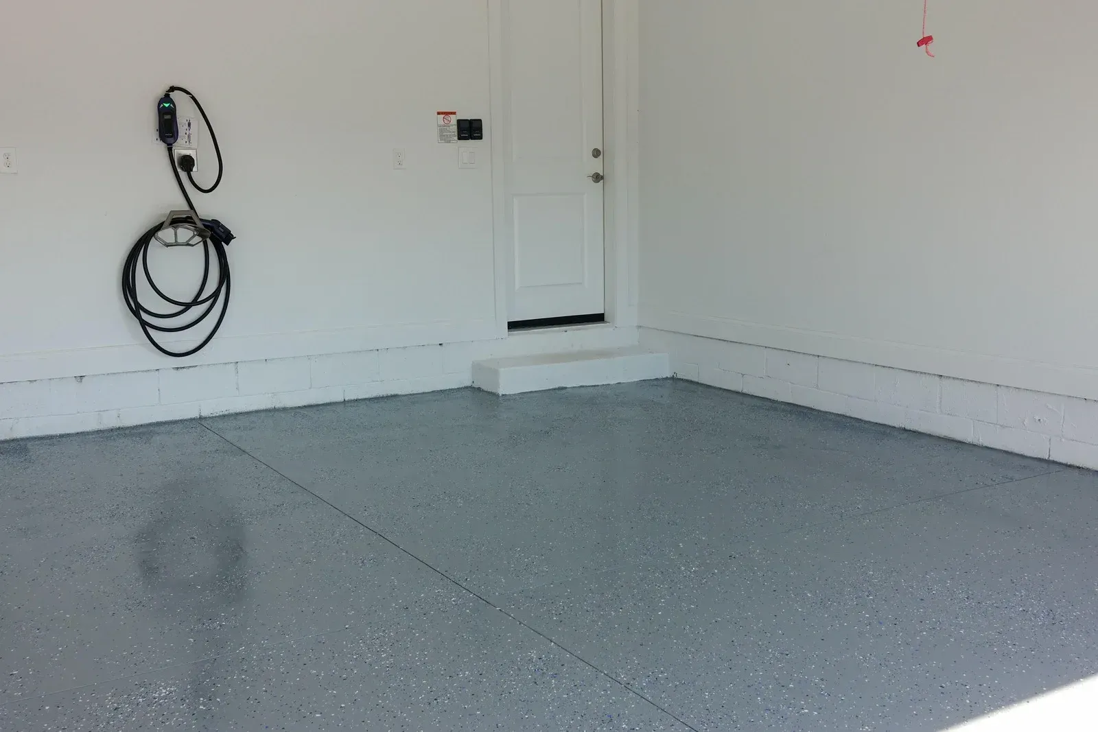 A garage with a gray speckled epoxy floor, a white door, and an electric vehicle charging cable coiled on the wall.