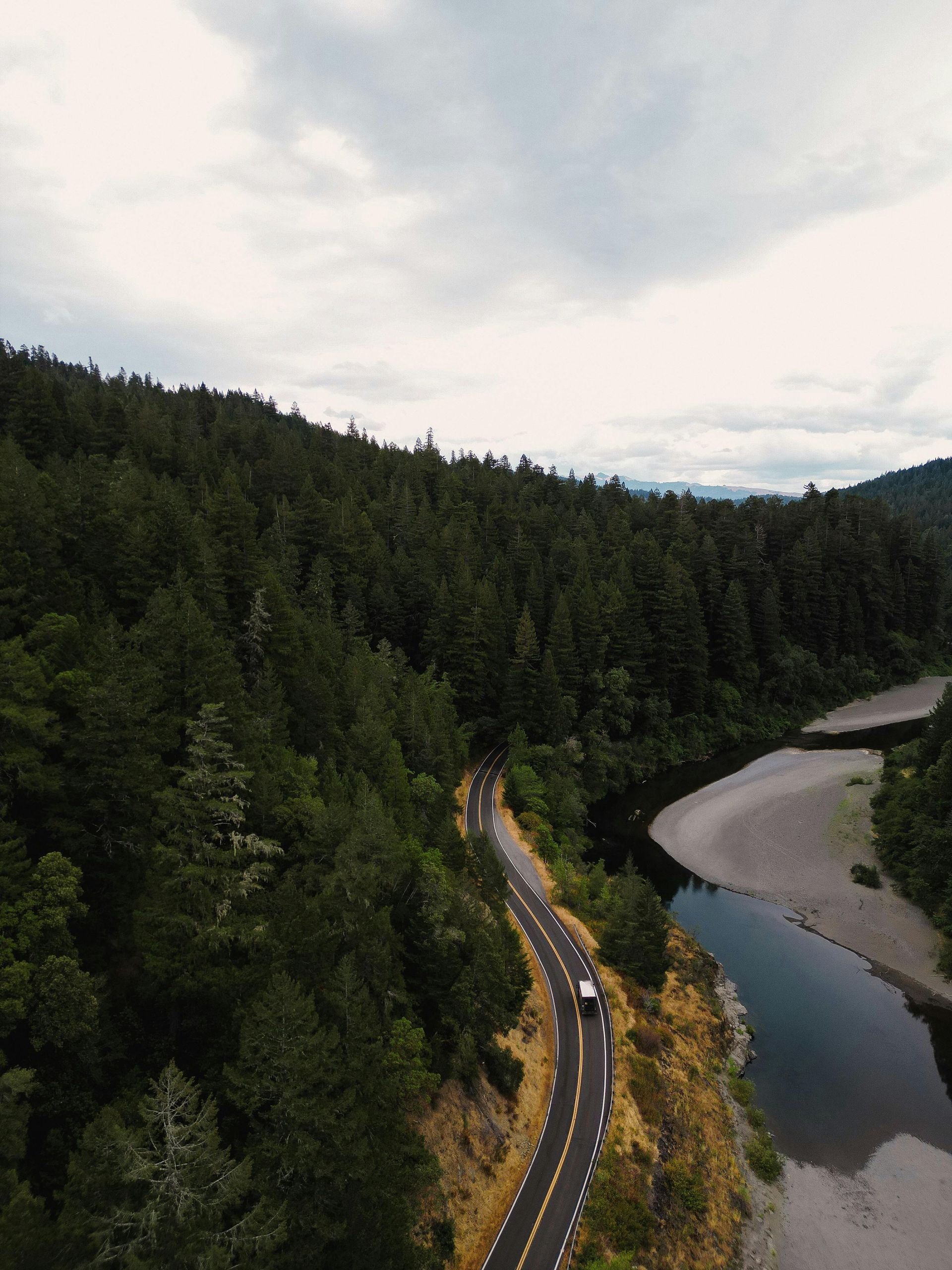Winding road through lush green forest alongside a river; cloudy sky.