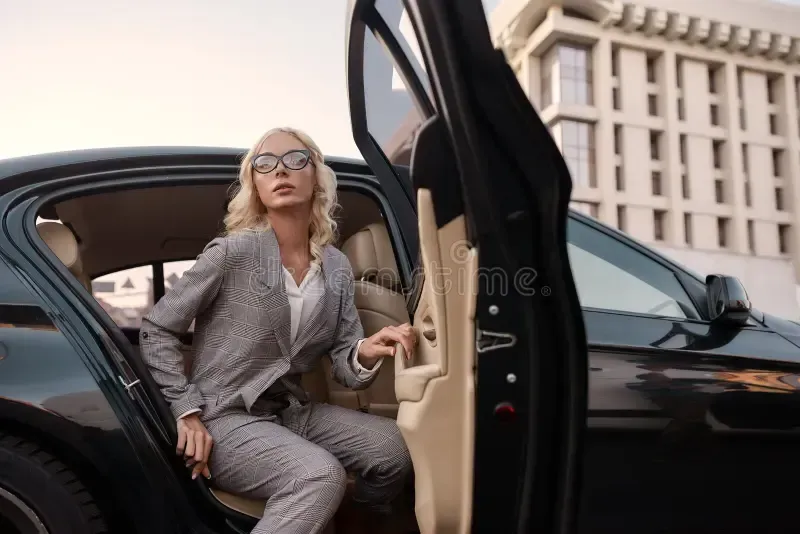 Blonde woman in business suit exiting black car, holding the door open, looking ahead.